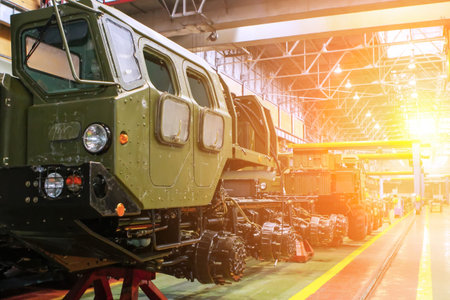 Military vehicles are lined up inside a spacious industrial facility, illuminated by warm sunset...