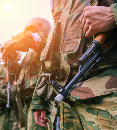 Groups of soldiers in camouflage gear stand ready for training, holding rifles as the sun shines...