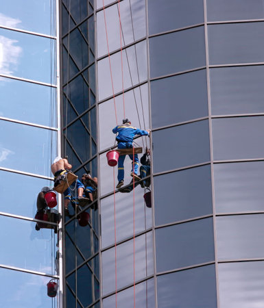 Two workers are using ropes to clean the glass windows of a tall building in the city center while...