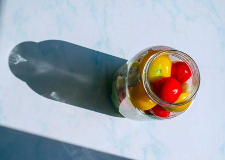 A glass jar holds vibrant red, yellow, and green cherry tomatoes on a marble surface, illuminated by sunlight.の写真素材