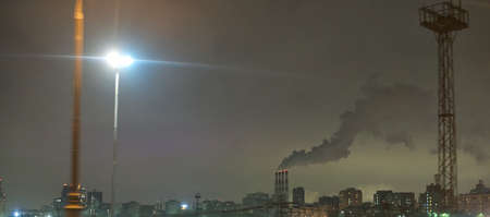 In the distance there are large pipes of a thermal power plant from which smoke is coming down. Late night. There are some buildings around. Sad urban landscapeの写真素材