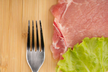 A piece of raw red meat with a lettuce leaf and a fork on a cutting board. Cooking steak at home.の写真素材