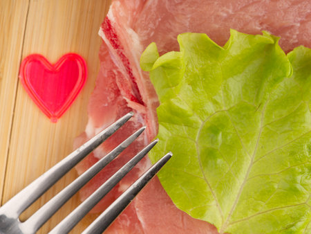 A piece of raw red meat with a lettuce leaf and a fork on a cutting board. A heart lies nearby.の写真素材
