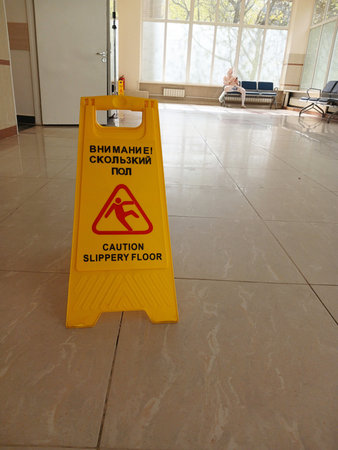 A sign cautiously wet the floor in the hospital lobby. In the background, a male patient is sitting on a bench. Vertical.の写真素材