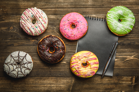 Multi-colored assortment of donuts with sprinkles and frosting on dark wooden background. with a notebook and penの写真素材