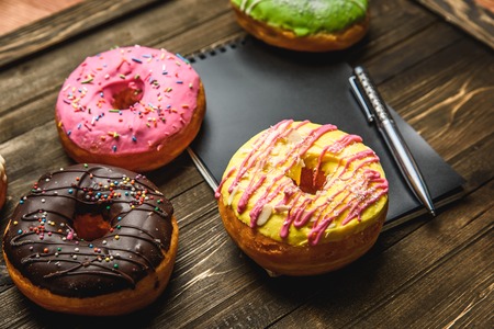 Multi-colored assortment of donuts with sprinkles and frosting on dark wooden background. with a notebook and penの写真素材
