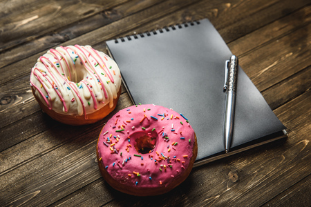 Multi-colored assortment of donuts with sprinkles and frosting on dark wooden background. with a notebook and penの写真素材