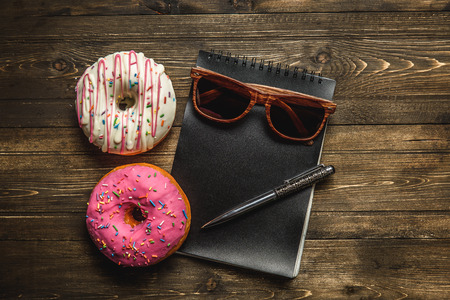 Multi-colored assortment of donuts with sprinkles and frosting on dark wooden background. with a notebook and pen and sunglassesの写真素材