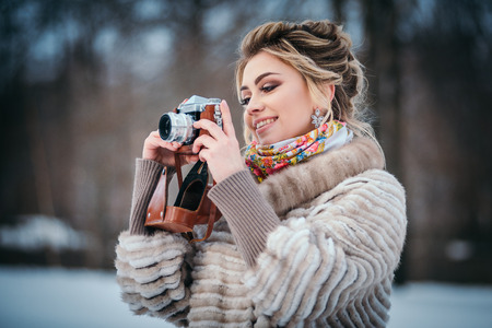 brunette girl is standing in a winter snow park, holding an old film camera and looking into the distanceの写真素材