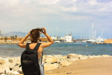Blonde-haired tourist girl with a black backpack goes along the waterfront, parking for yachts on the horizonの写真素材