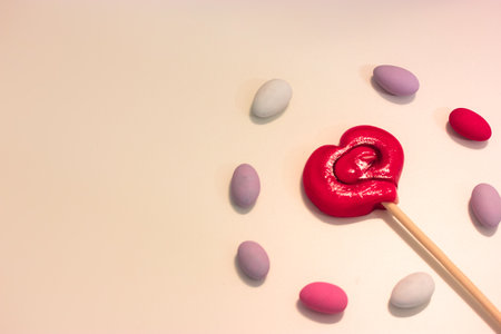 Lollipop on a stick in the shape of a red heart on a white table surrounded by colorful candy, layout. A sweet treat for Valentines Dayの写真素材