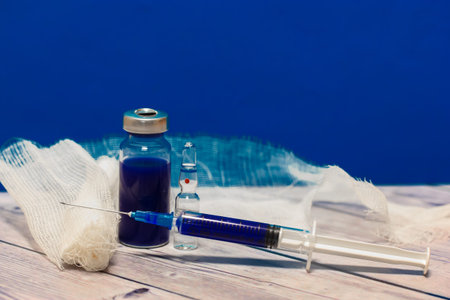 A glass medical transparent ampoule, syringe and bandage on a table against blue wall. pandemic...