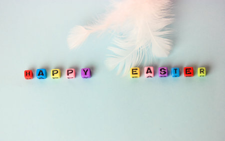 Happy Easter lettering text made of multicolored wooden letters and white feather on a soft blue...
