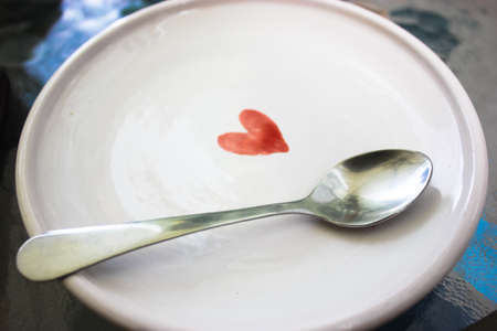 A white ceramic saucer with a picture of a red heart and a silver teaspoon on the surface of the plate. Clean utensils. High quality photo.の写真素材