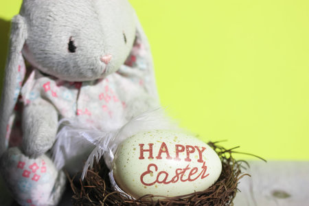 Toy bunny on yellow green background and a nest with an Easter egg inside. Egg with the inscription...
