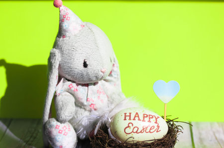 Toy bunny on green background and a nest with an Easter egg inside. Egg with the inscription Happy...
