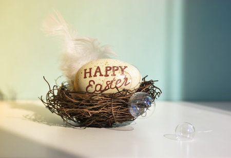 A nest with an Easter egg and white bird feather inside. Light blue background. The words Happy...