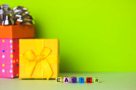 Happy Easter multicolored words written with wooden alphabet letters on a bright green background. Gift boxes in the background. Greeting card for Christian or Catholic Easter. Place for text. Flatly.の写真素材