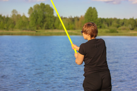Fisherwoman with a yellow fishing rod on a background of a pond and a blue sky on a summer sunny...