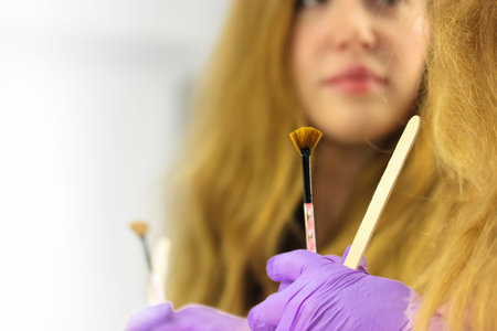 A red-haired girl holds a wooden disposable cosmetic spatula for applying cream and a brush for masks. Tools for a beautician's work. Skin care for the face and body. White background, place for text.の写真素材