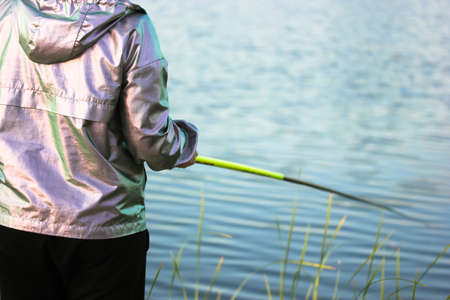 Fisherwoman with a yellow fishing rod on a background of a pond, river, lake on a sunny summer day. Woman catching fish. Active leisure and recreation in nature reserve. Angling on a bank. Blue water.の写真素材