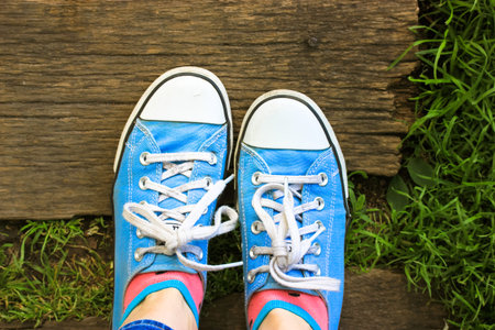 A girl in blue sneakers stands on a wooden bridge. Top view, flatly of women's feet on green young...