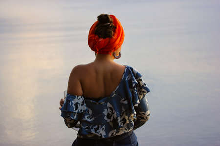 Back view of a woman in a one-shoulder blouse with an orange turban sitting by the sea on a summer evening, twilight. Glaze on a blue water. Rest by the ocean, tranquility, harmony, calmness concept.の写真素材
