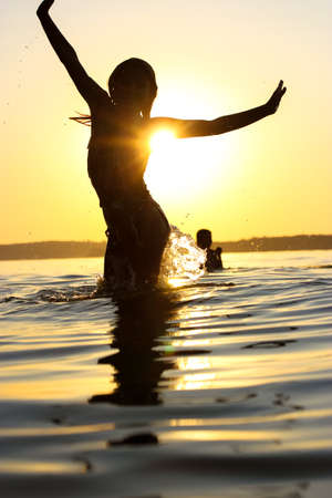 Silhouette of happy children playing in a water. Kids frolic, have fun, jump in a calm water of the sea, lake, river at sunset. Summer vacations, holidays on the coast. Happy childhood. Splash water.の写真素材