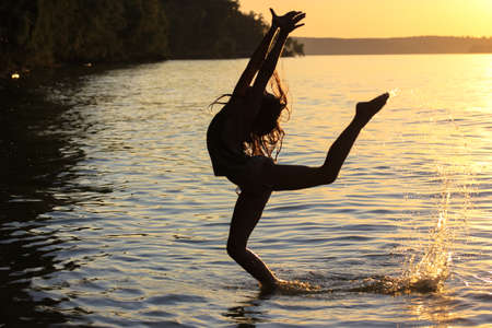 A professional gymnast girl is engaged in outdoor sports on a beach on the sunset. A teenage doing stretching exercises on seashore. An elegant silhouette of a girl. Splash water of a sea, lake, riverの写真素材