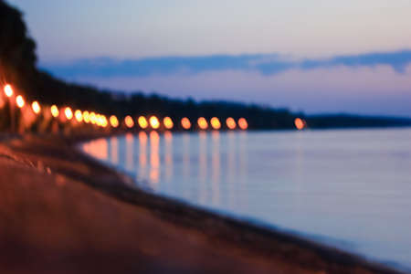 Blurred evening promenade by the sea in defocus. Yellow lantern lights on lampposts and blue calm sea, river at dusk. Night descends on a city. A strong blur. Beautiful seascape abstract background.の写真素材