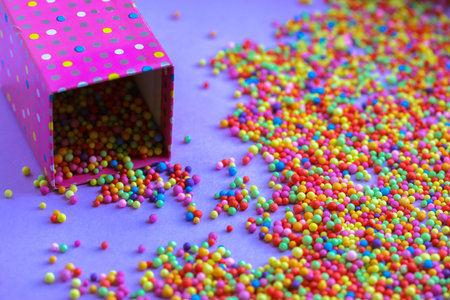 A pink polka dot gift box flatly, lots of multi-colored foam beads of various colors scattered on a...