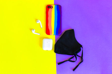 Black medical face mask, purple sunglasses, wireless headphones, phone in a multi-colored striped case on yellow-violet background flatly. New normal. Pandemic . Accessories for summer vacation.の写真素材