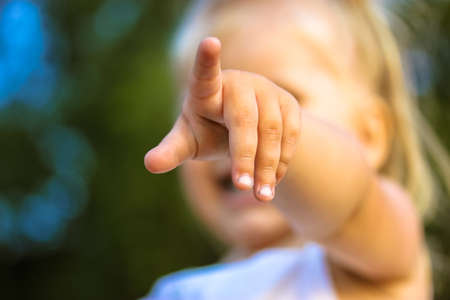 A little blonde infant girl pointing finger into a distance straight ahead. Baby girl's hand in focus. Age 2-3 years old. Young children in a green park blurry background at summer day. Cute child.の写真素材