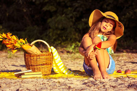 Beautiful teenage girl in a hat with a watermelon lollipop sits on a sandy beach at sunny summer day. Cute female kid on a picnic. Wicker basket, baguette, flowers, peaches, books. Holidays, vacation.の写真素材