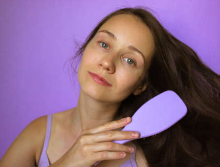 Portrait of pretty smiling girl, holding a purple comb and brushing her long natural brown hair. Self care, beauty concept. Woman 25-30 years old standing isolated on violet background. Space for textの写真素材