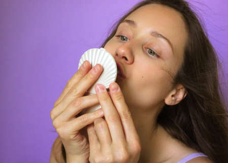 A young, pretty cute brunette woman holding a pearl pocket mirror and kissing her reflection in it. Narcissism, self-love, selfishness, I love me concept. A girl in a purple background. Beauty day.の写真素材