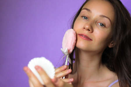 A girl smiling stroking facial skin with a pink fluffy make-up powder puff. Happy young dark haired woman applying make-up on her face, admiring herself in a pocket mirror. Beauty concept. Copy space.の写真素材