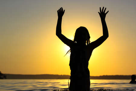 Silhouette of happy teenage girl standing a confident pose in a river, pond, lake water at sunset with hands in the air, fingers keeping apart. Summer vacations, holidays on a coast. Happy childhood.の写真素材