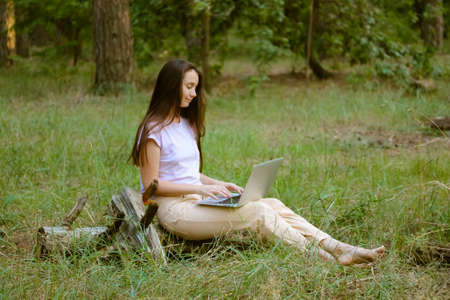 A beautiful young woman, girl with long brown hair sitting in a clearing in a woods, green forest, working at laptop. Remote work, study. Student with a computer in nature. Internet. Cinematic filter.の写真素材