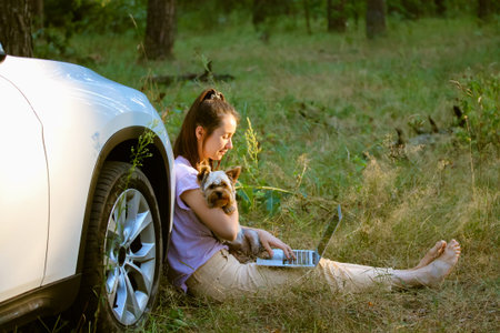 A young woman, girl with a small dog Yorkshire terrier sitting near a white SUV car in forest,...