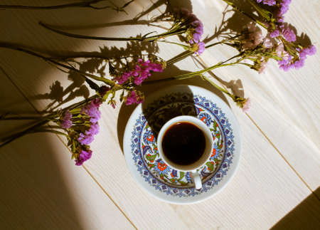 Turkish coffee in traditional blue porcelain cup at saucer on a white wooden floor top view. Contrasting shadows. Hot espresso drink in a morning light flatly. A composition of dried purple flowers.の写真素材