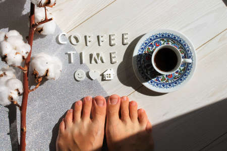 Time for a black coffee. Woman's legs, feet on a white wooden floor. Espresso in a cup standing on a white wooden floor flat lay, top view. Contrasting shadows in early morning. Cotton flower branchの写真素材