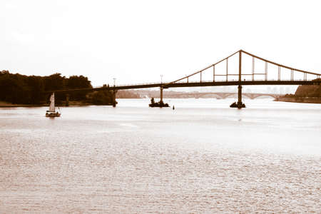 Kiev, Ukraine. July 1, 2021. Pedestrian bridge over a Dnieper River in Kiev. Beautiful scenery postcard evening dusk view of a sailboat floating by the water and bridges on a horizon. Brown toning.のeditorial素材