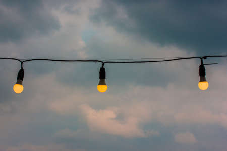 Decorative Edison lamp garland hanging outdoors against a blue overcast stormy sky at the evening. Artificial warm yellow light, electricity concept. Bulb lamps on a street. Black electrical wire.の写真素材