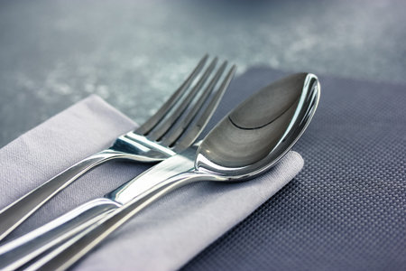 The soup spoon and metal fork lie on a gray napkin on a black gray surface top view. Table setting,...
