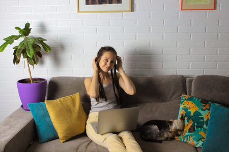 Pretty young smiling girl 20-30 years old sitting on a gray couch with a laptop next to a small sleeping dog. Studying, working remotely from home in modern interior. Cozy atmosphere. Stay at home.の写真素材