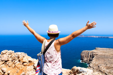 A tourist with a straw hat standing on a hill with her hands up to greet a blue sea at sunny summer day. Female traveler view from a back. Rest on a beach, the ocean. Travel the World. Explore Earth.の写真素材