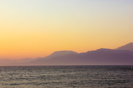 Seascape with silhouettes of hills and mountain. Warm sunlight breaking from behind the mountains at...