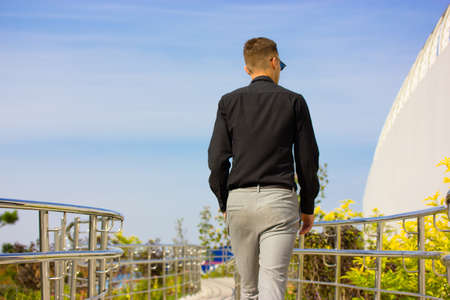 A businessman in business suit, black shirt, gray pants, sunglasses walks along a winding path in a city park. A man from the back against blue sky. Metal fence on a flowerbed. Urban people lifestyle.の写真素材