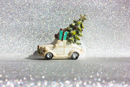 White toy truck car with green Christmas pine tree on roof rides on a silvered shining background. Minimalistic happy New Year's Eve background. Greeting card, copy space. Blue gift box delivery.の写真素材